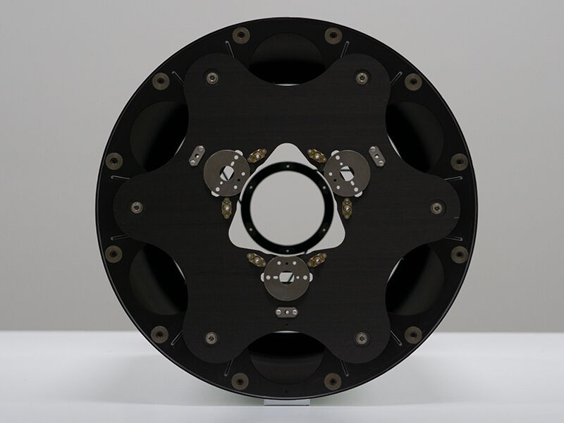 Satellite imaging optical metering structure. Image shows circular side panel, which is black carbon fiber side panel with metallic inserts. The structure is laying on a white tabletop with a white background. 